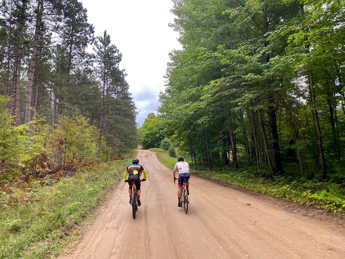 Two cyclists on a long and straight gravel road in the woods.